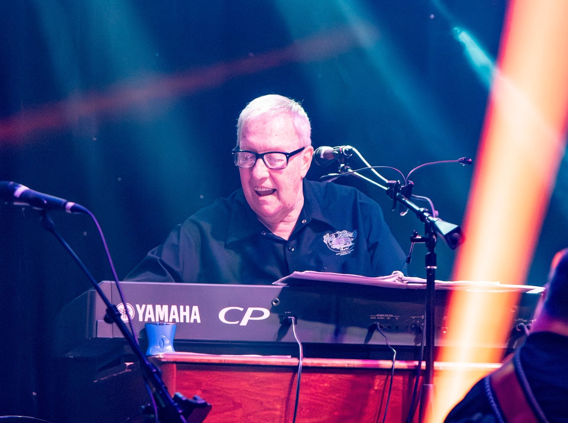 A person performing at a keyboard on a dimly lit stage, speaking into a microphone with a wooden piano base visible.