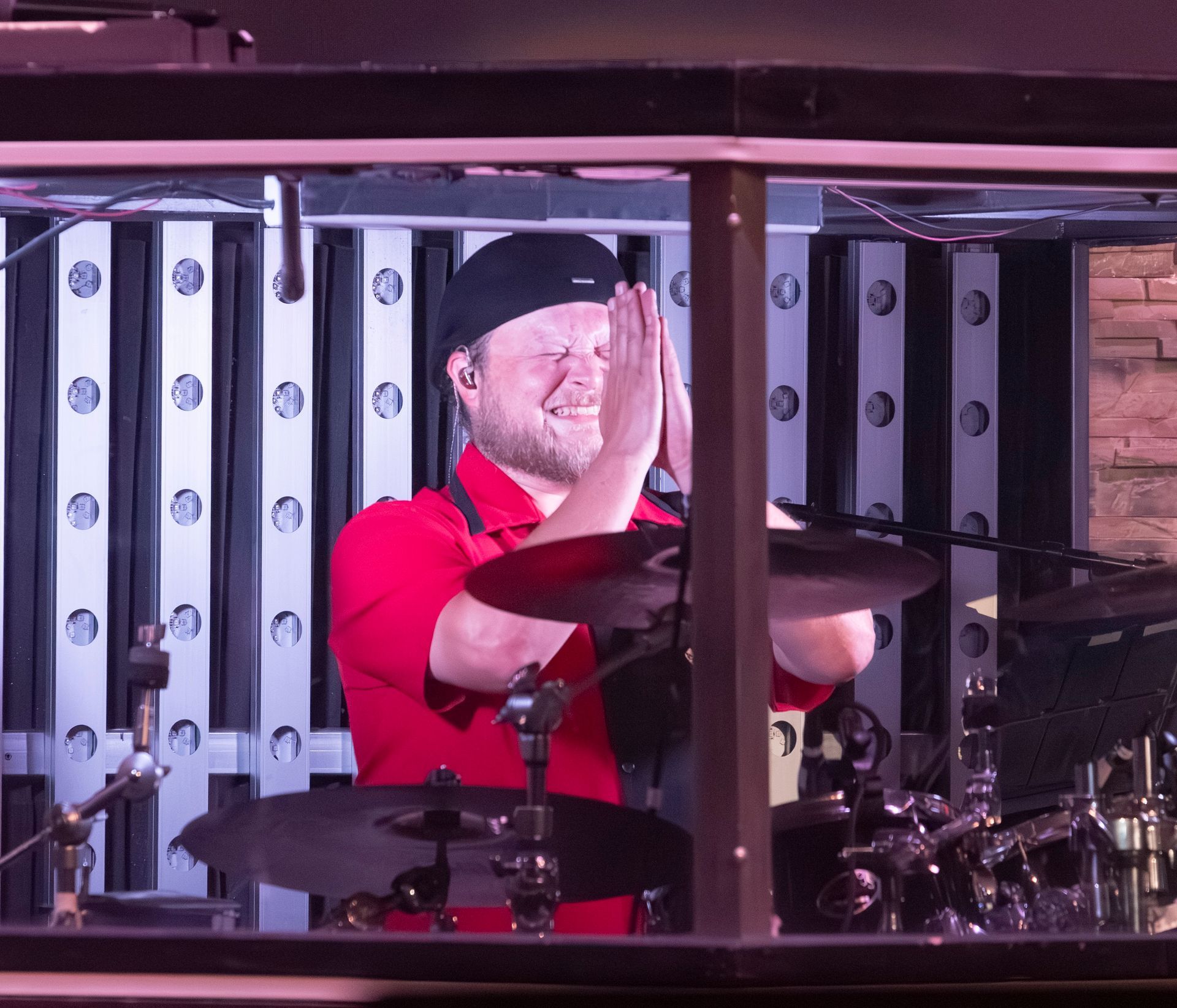 A drummer wearing a red and black shirt points upward while sitting behind a drum kit in a dimly lit studio setting.