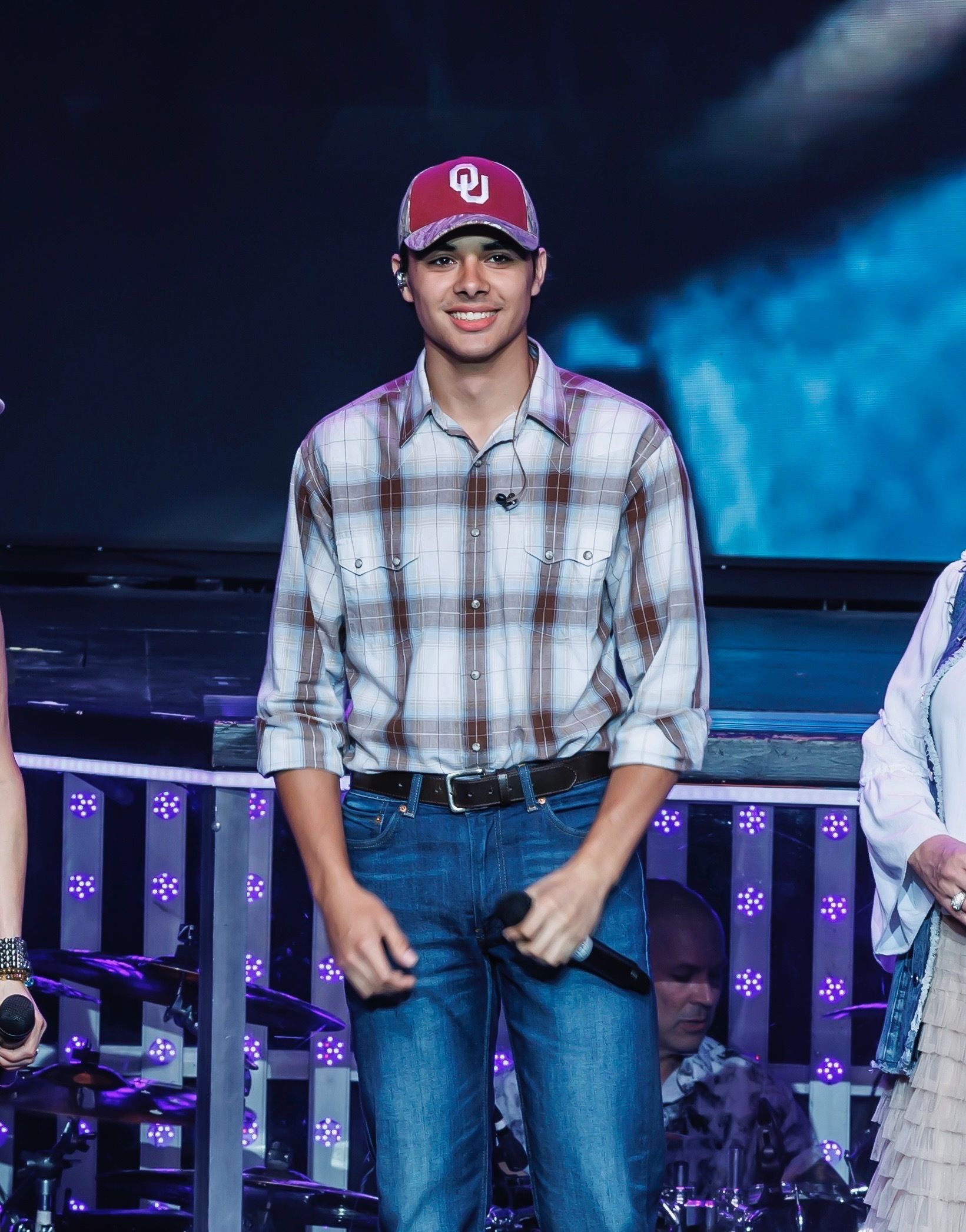 A person stands on a stage wearing an OU baseball cap, a plaid button-down shirt, and blue jeans, holding a microphone.