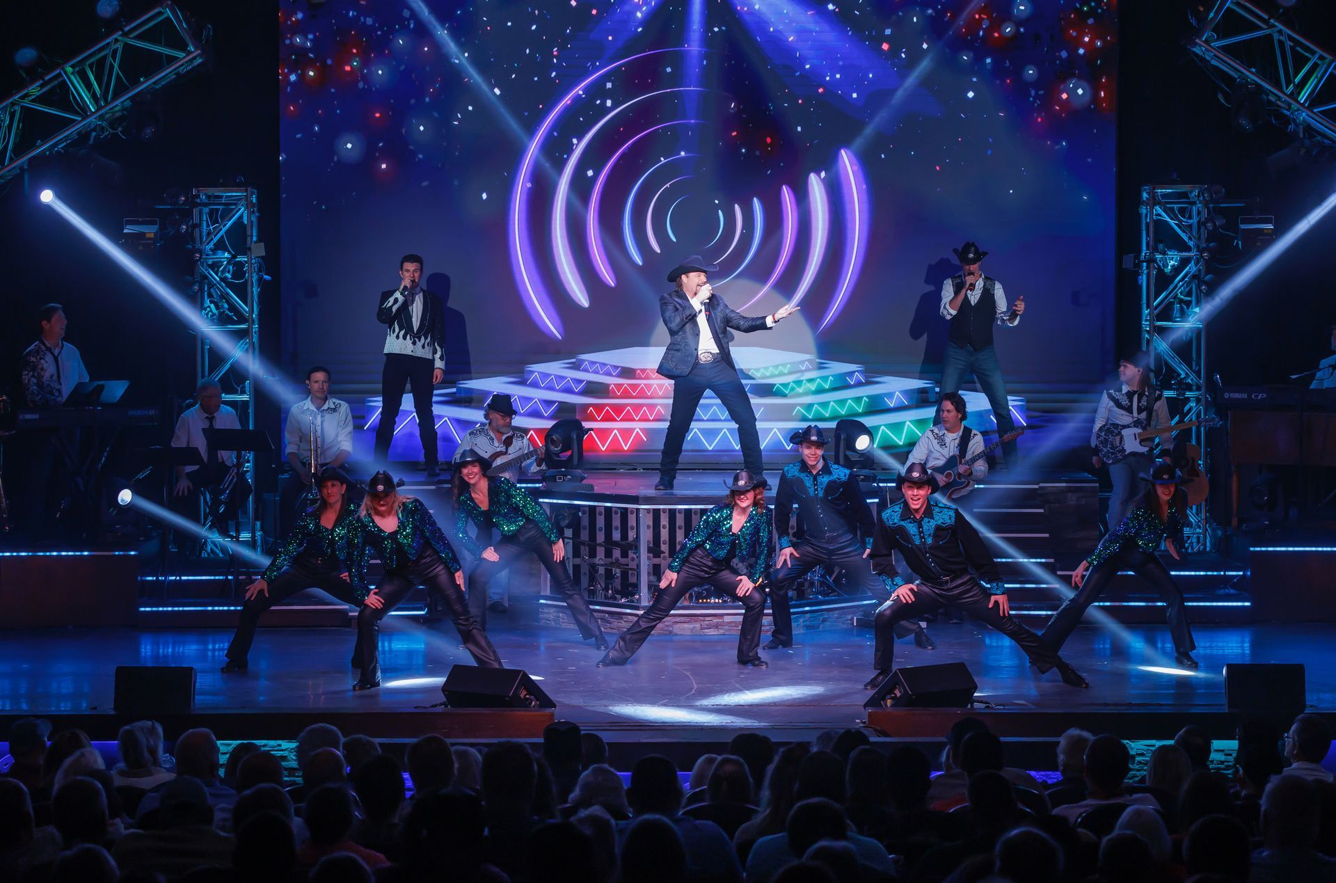 A performer sings center stage, surrounded by backup dancers in green outfits, set against a vibrant, illuminated backdrop.