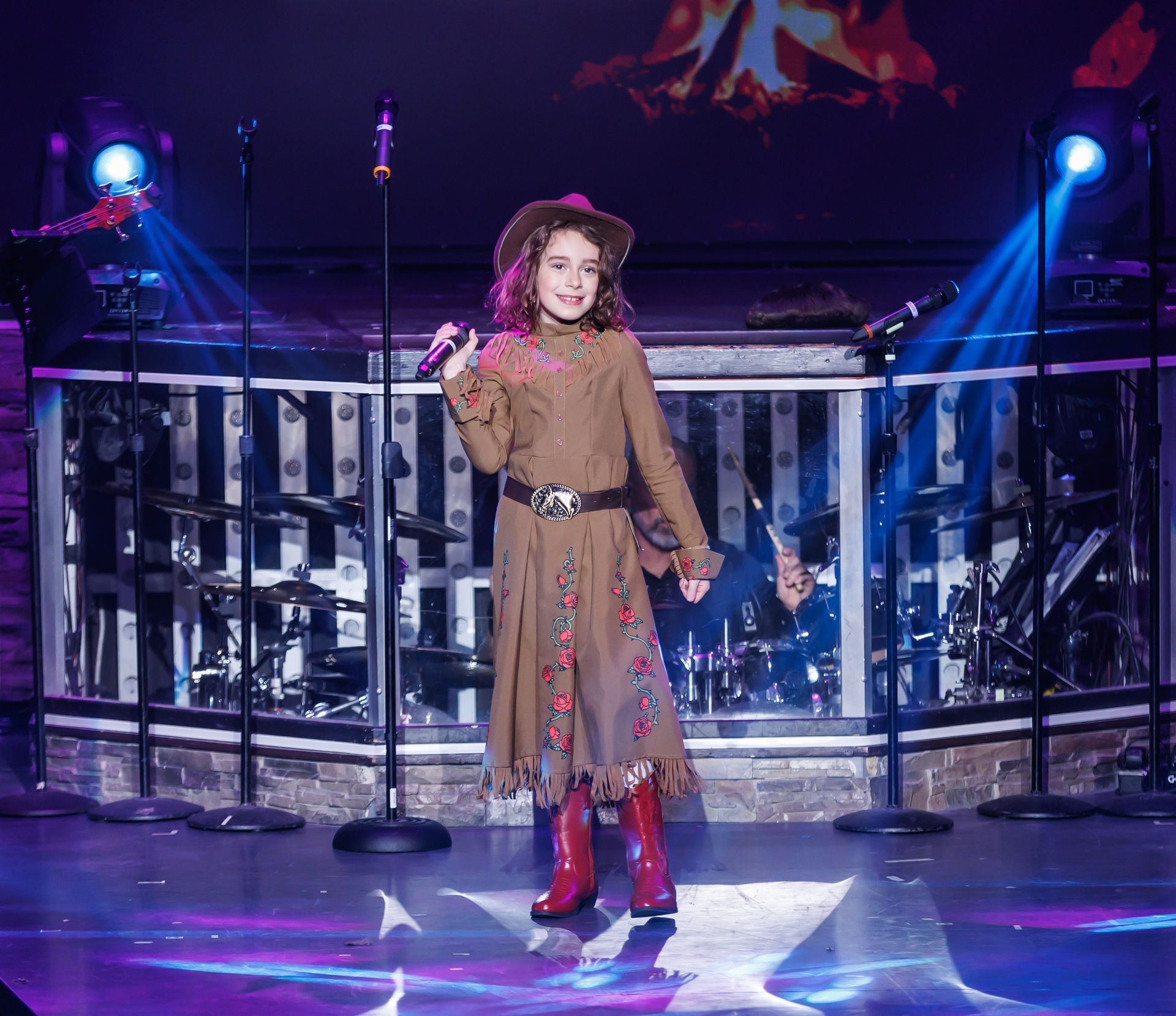 A person in a brown Western-style dress and red cowboy boots sings into a microphone on a stage with bright blue lights.