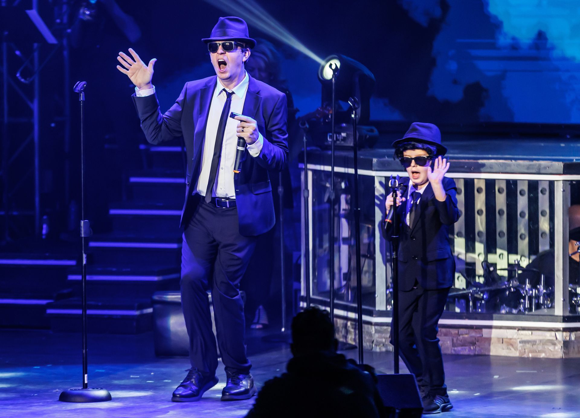 Two performers in suits, ties, and fedoras singing into microphones on a stage with blue lighting.
