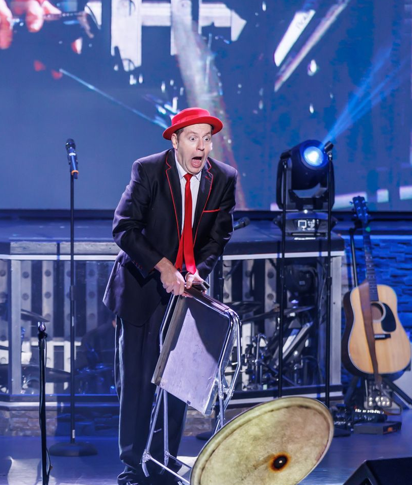 A performer in a black suit and red hat stands on a stage with a shocked expression, holding a suitcase near a cymbal.