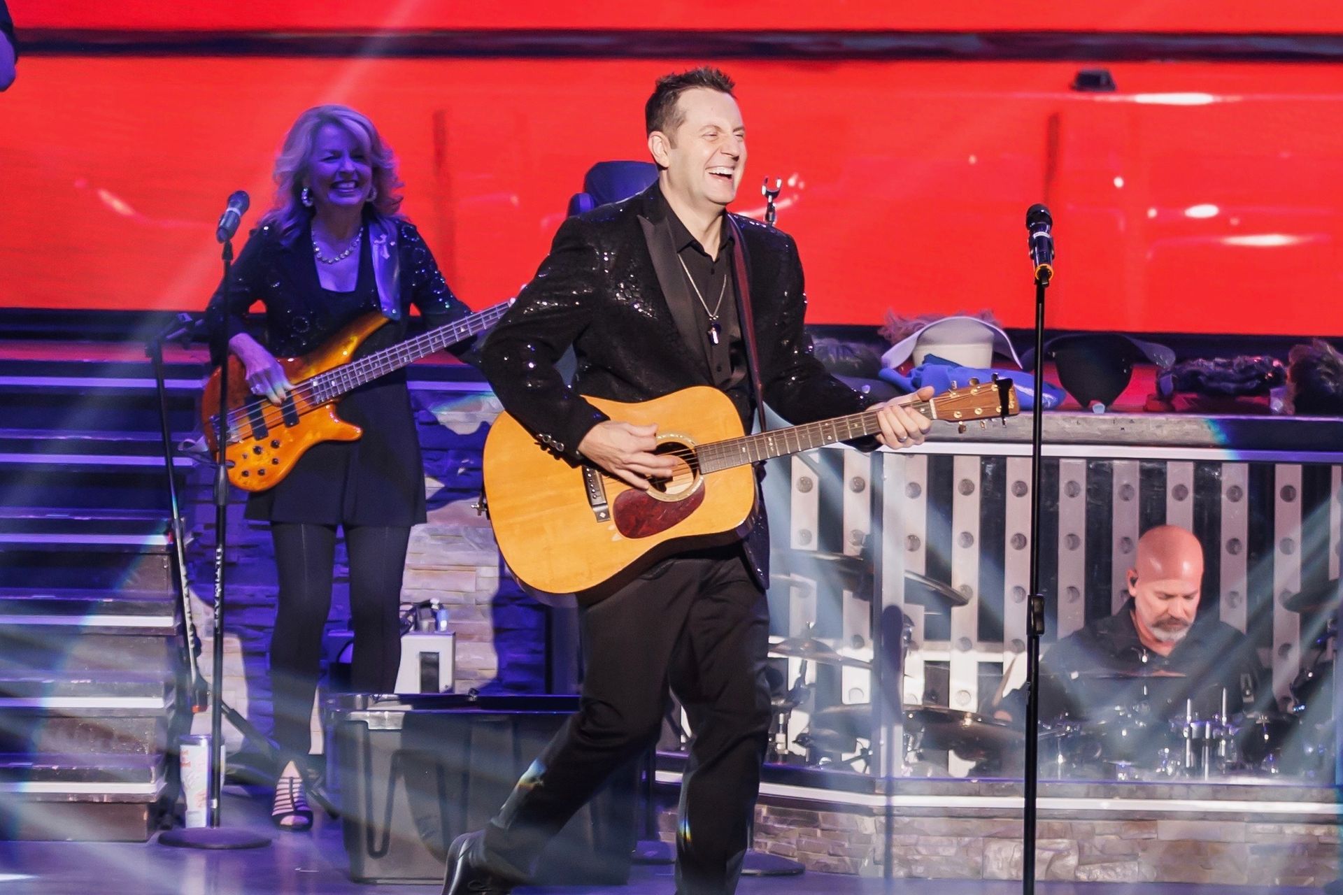 A musician in a sequined jacket plays acoustic guitar on a stage with a red background, backed by a bassist and drummer.