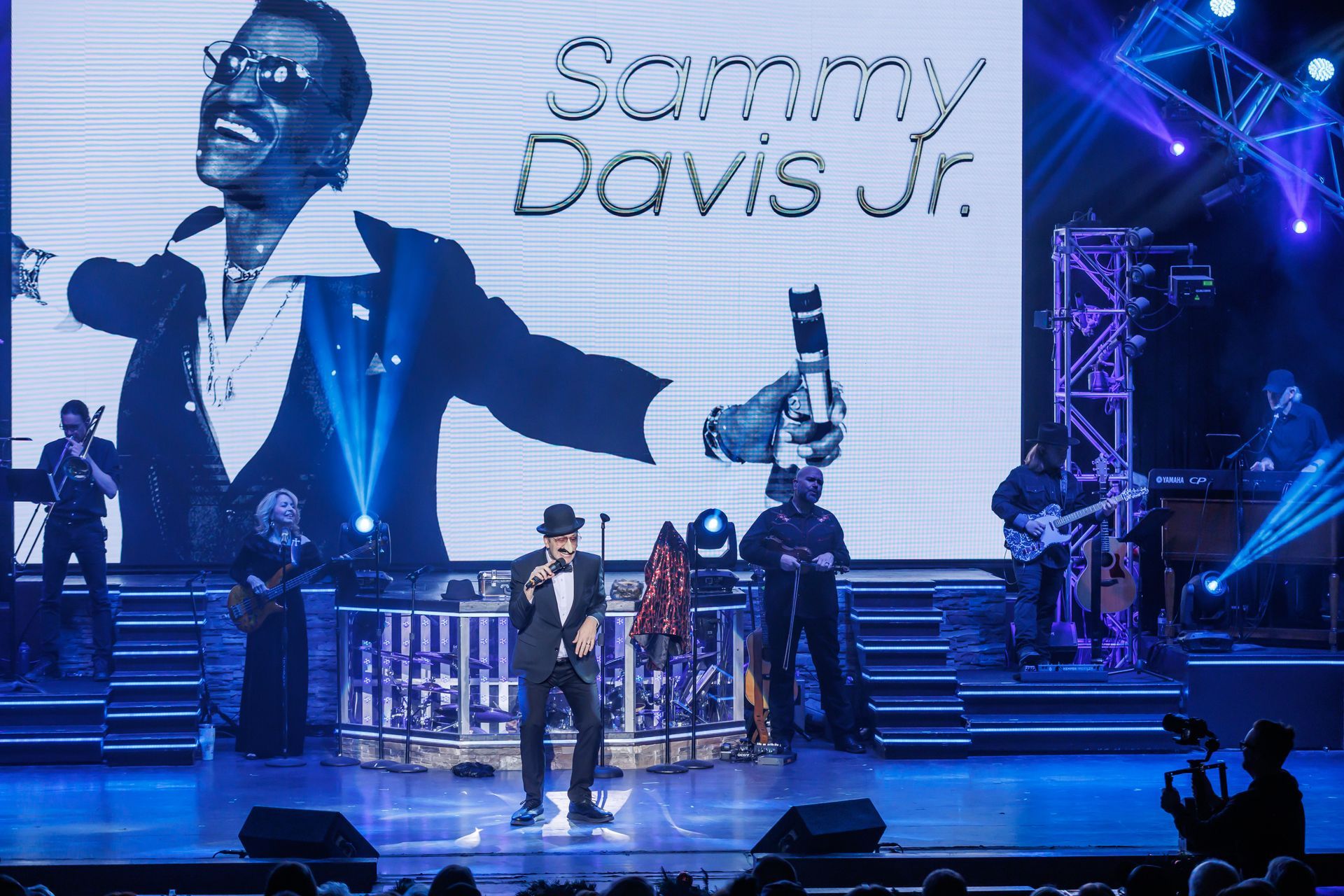 A performer in a suit and hat sings on stage in front of a large screen displaying Sammy Davis Jr. with a blue spotlight.