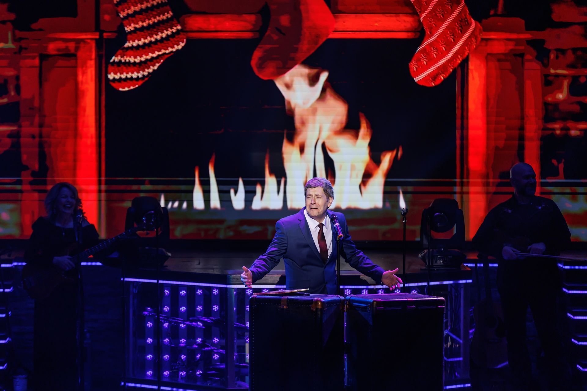 A person in a suit stands on a stage with a digital fireplace background and Christmas stockings, arms spread wide.