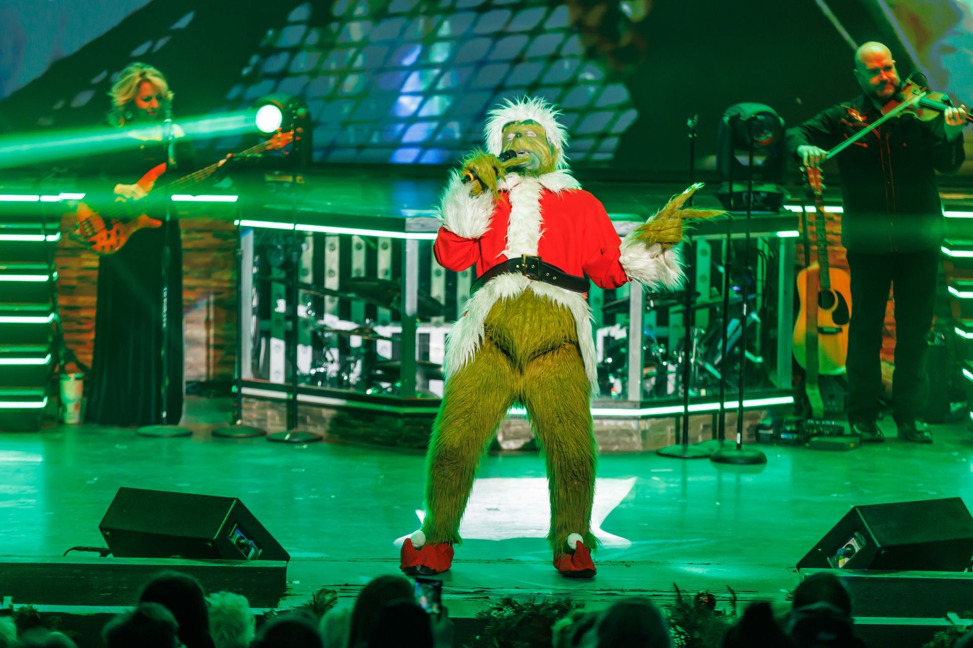 The Grinch, in a red Santa suit, sings into a microphone on a stage with a band under green spotlights.