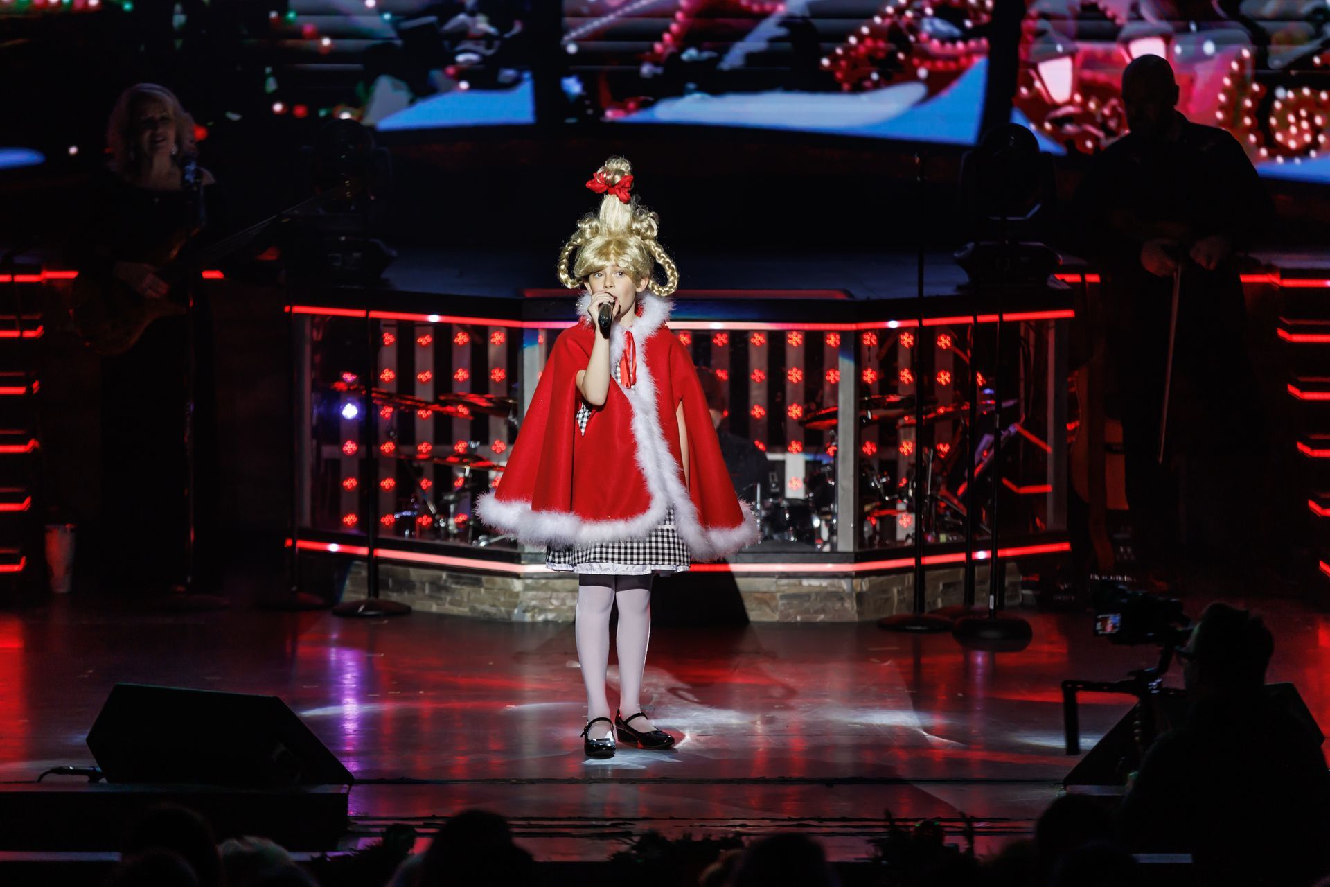A young performer dressed as Cindy Lou Who, wearing a red cape and elaborate blonde hairstyle, sings on a stage.