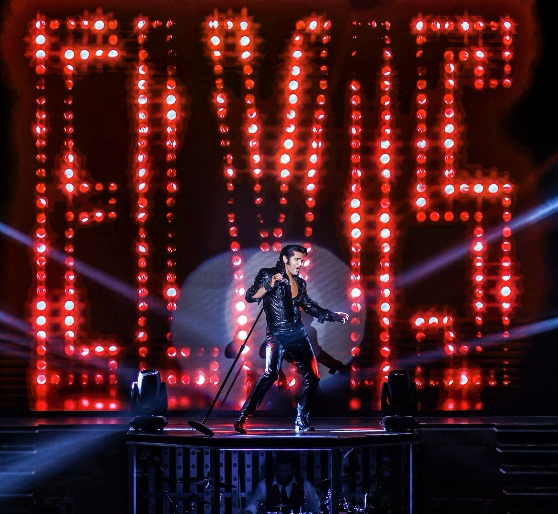 A person dressed in a black leather outfit performs on stage in front of a giant, illuminated red 