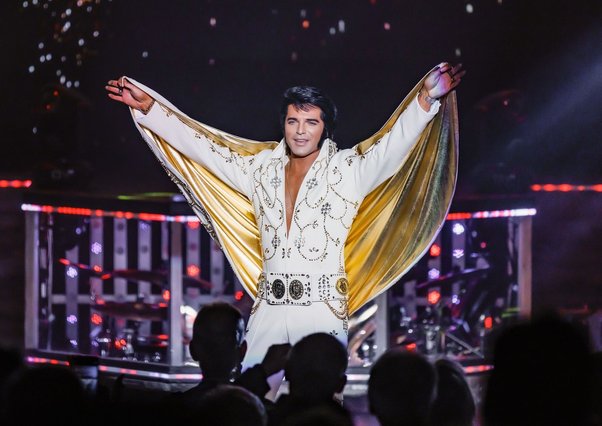 A performer in a black suit singing on stage with bright red lights, blue spotlights, and a background banner reading ELVIS.