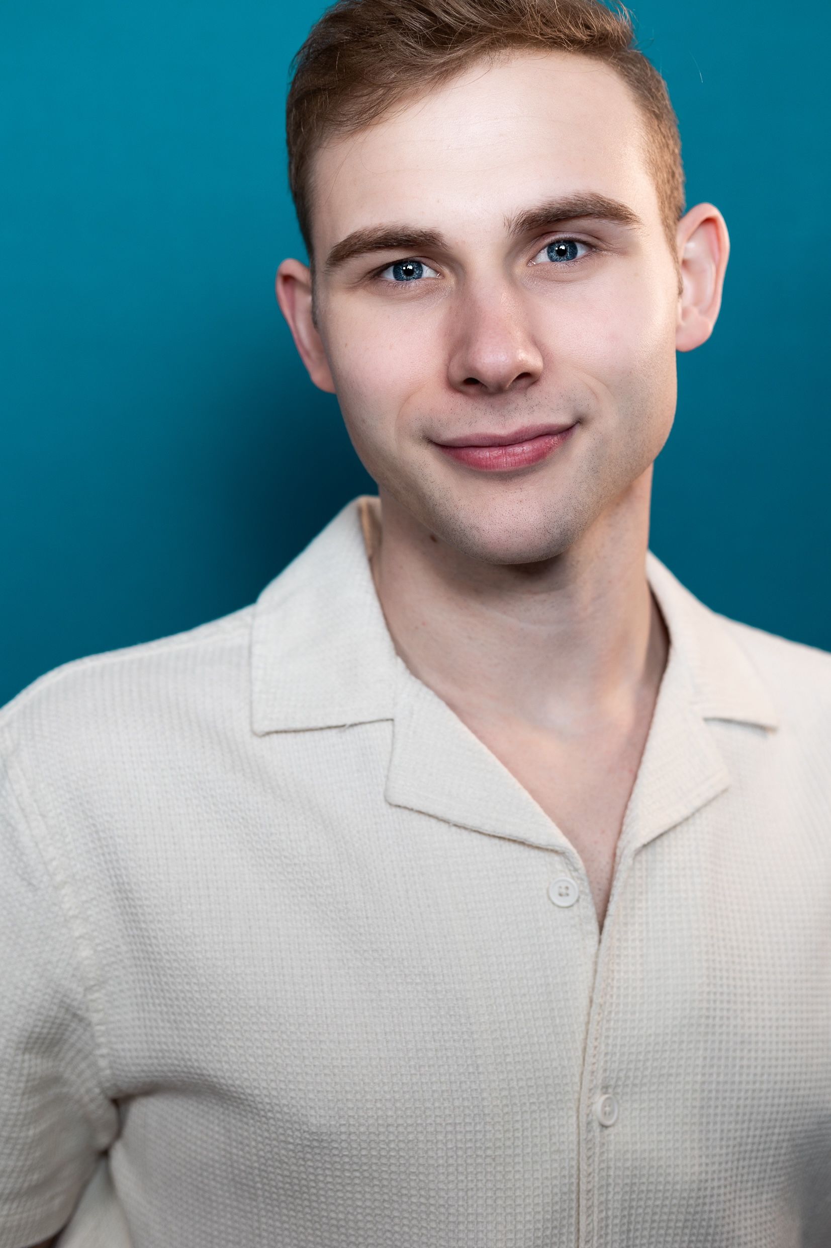 A person with short, light hair wears a beige button-down shirt against a solid teal background.