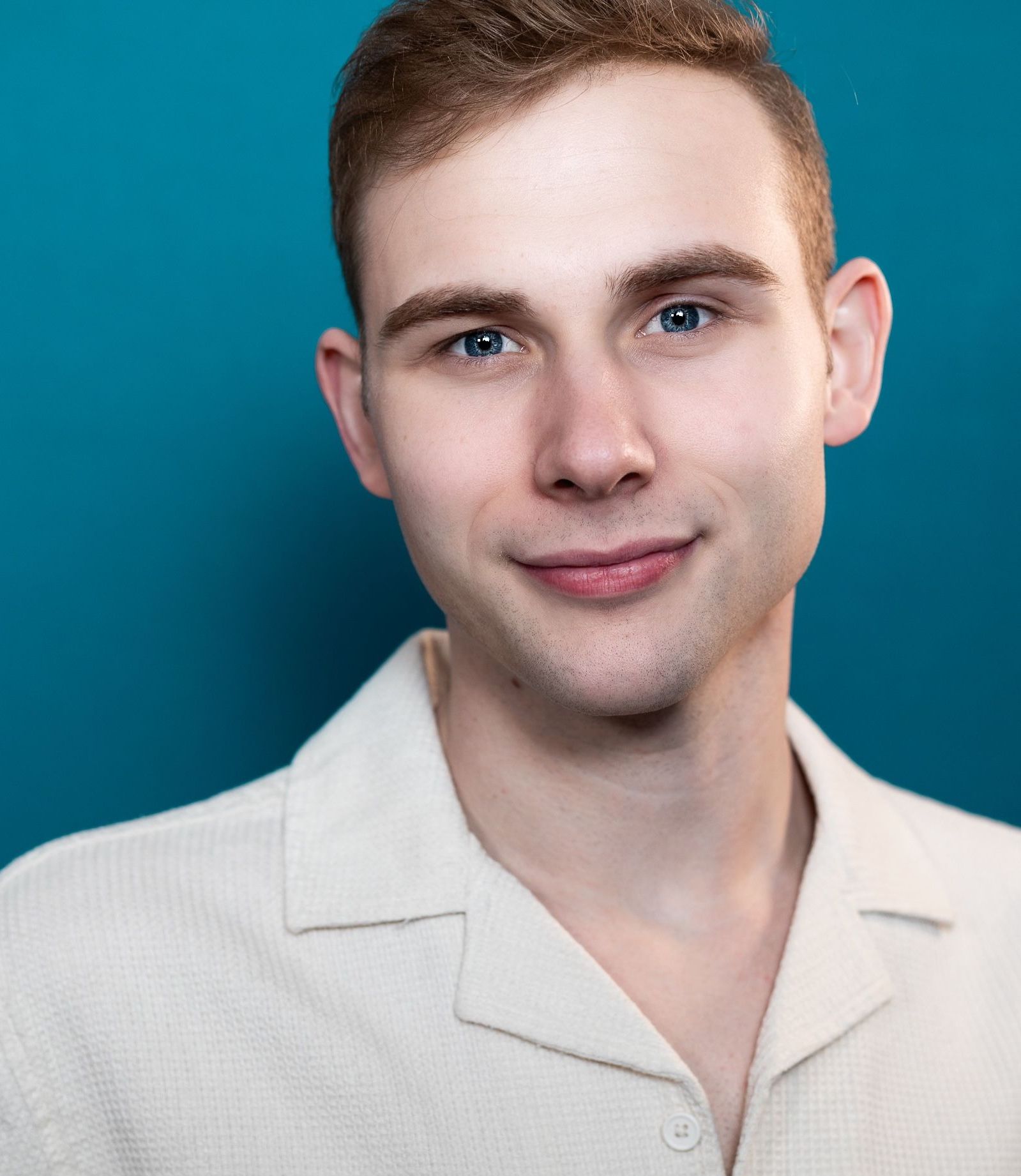 A person with short light hair wearing a cream-colored collared shirt against a plain teal background.