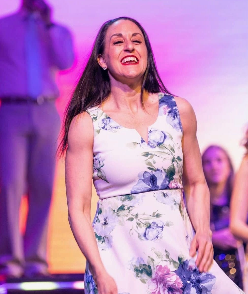 A smiling person in a floral dress stands on a stage during an event.