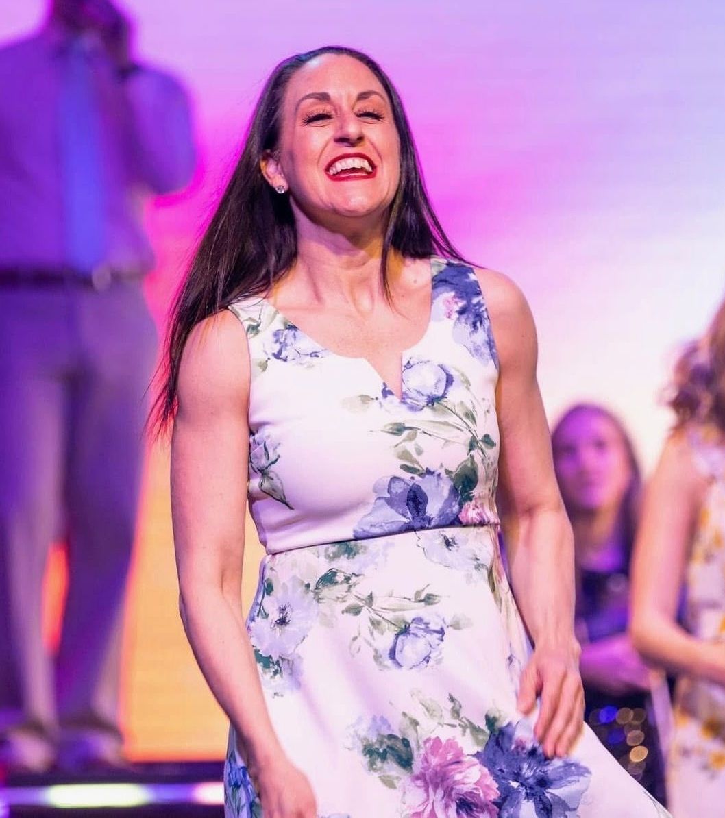 A smiling woman in a floral dress stands on a brightly lit stage in front of a blurred audience.