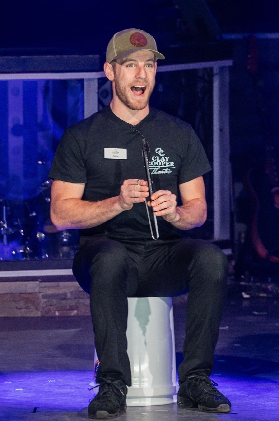 A person in a black shirt and baseball cap sits on a stool, smiling while holding a small wind instrument on a stage.