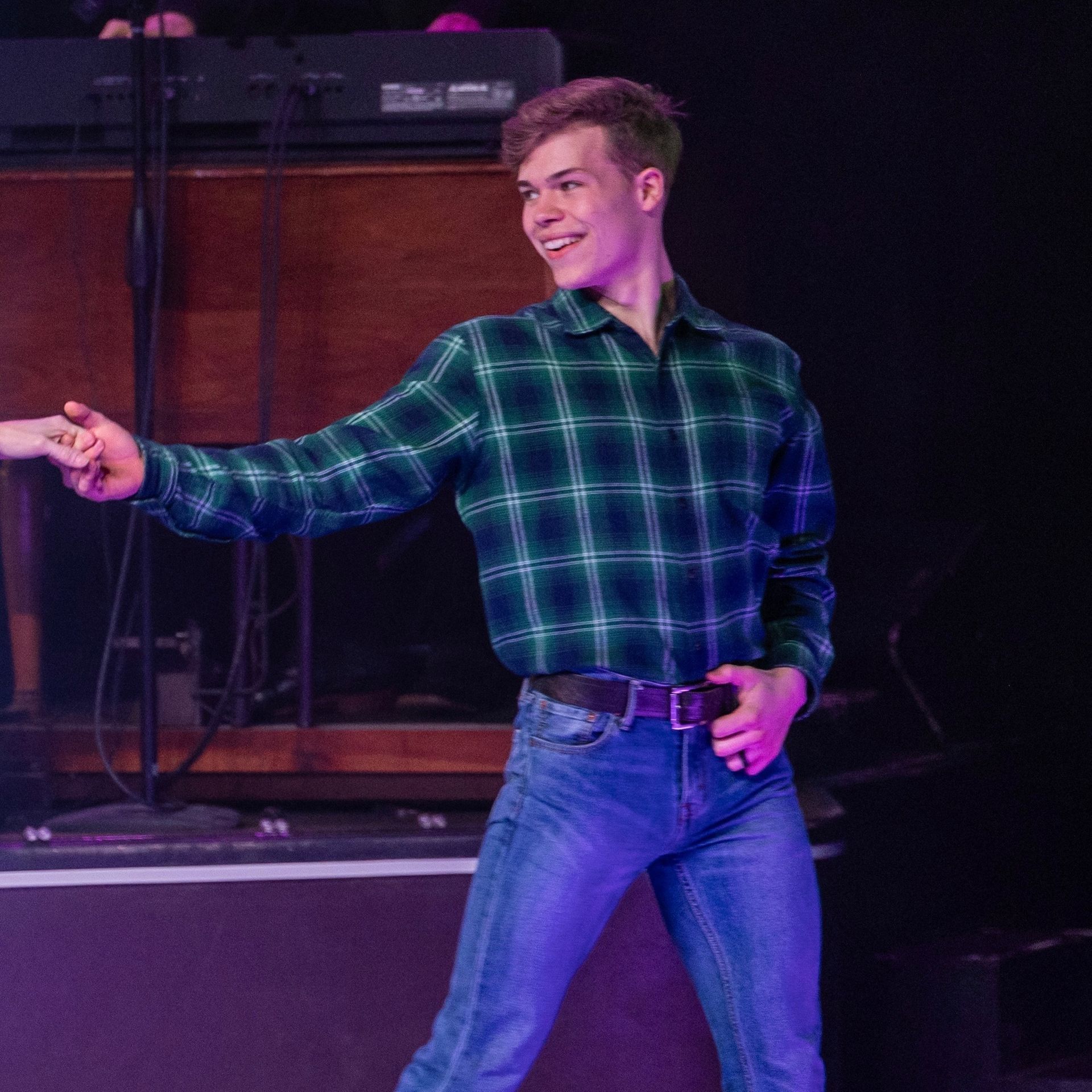 A person in a green plaid shirt and blue jeans smiles and reaches out with their right hand on a dimly lit stage.