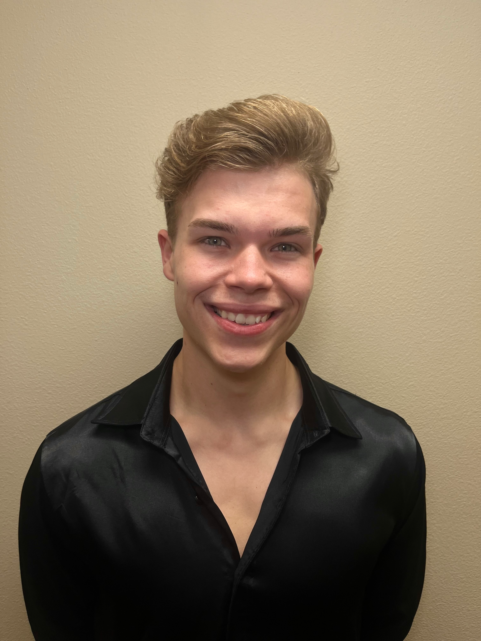 A smiling person with light brown hair in a styled quiff, wearing a black collared shirt against a beige background.