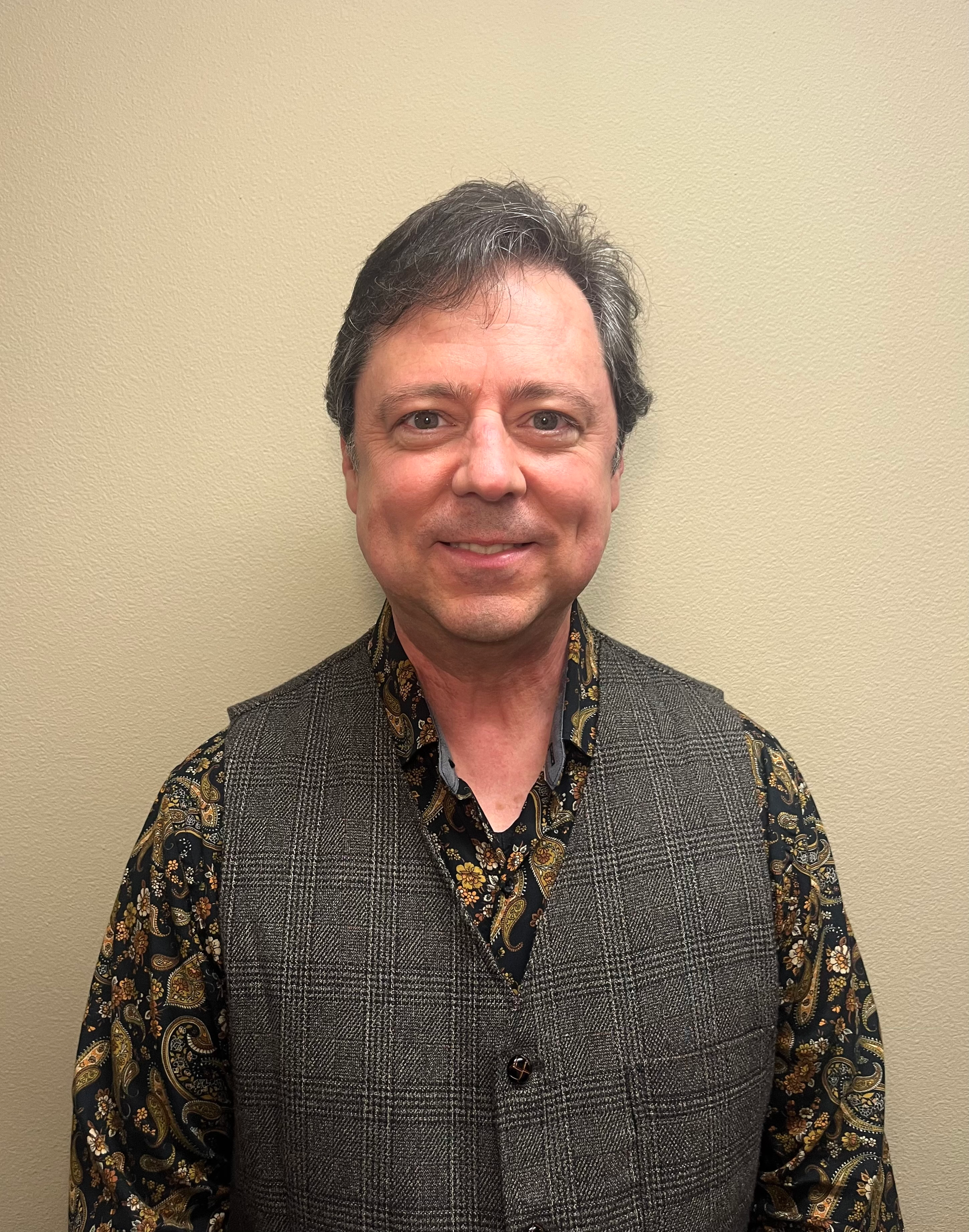 A person wearing a dark patterned shirt and a textured gray vest stands against a plain light-colored wall.