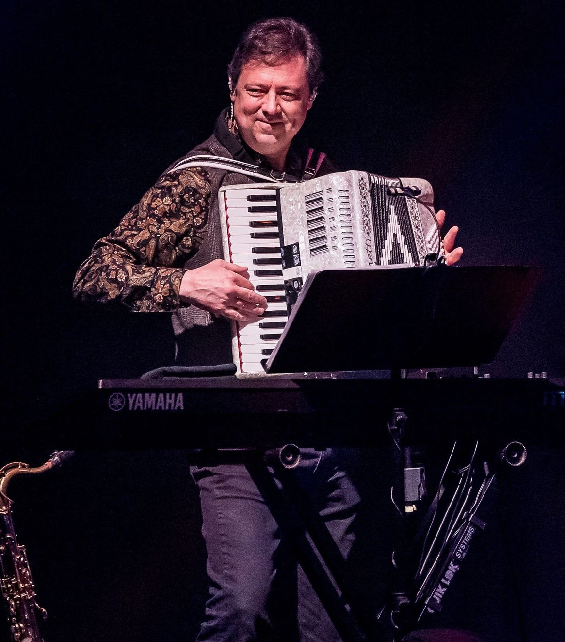 A performer plays an accordion while standing behind a Yamaha keyboard on a dark stage.