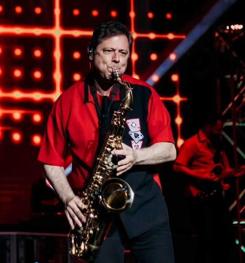 A performer plays a saxophone on stage against a backdrop of red, grid-patterned lights.