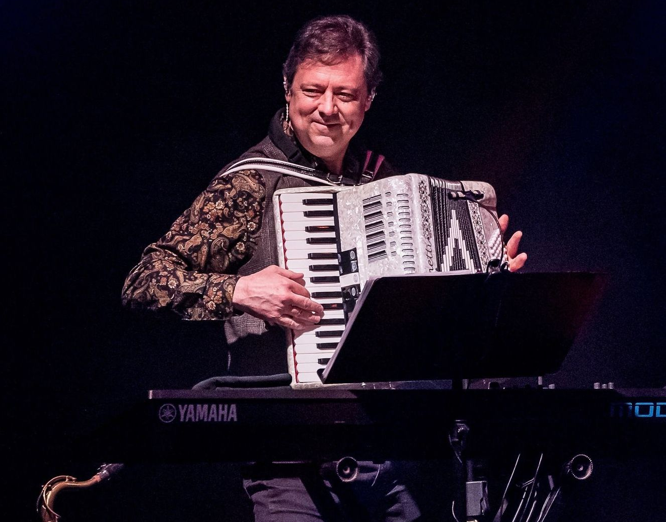 A musician playing an accordion in front of a Yamaha keyboard on a dimly lit stage.