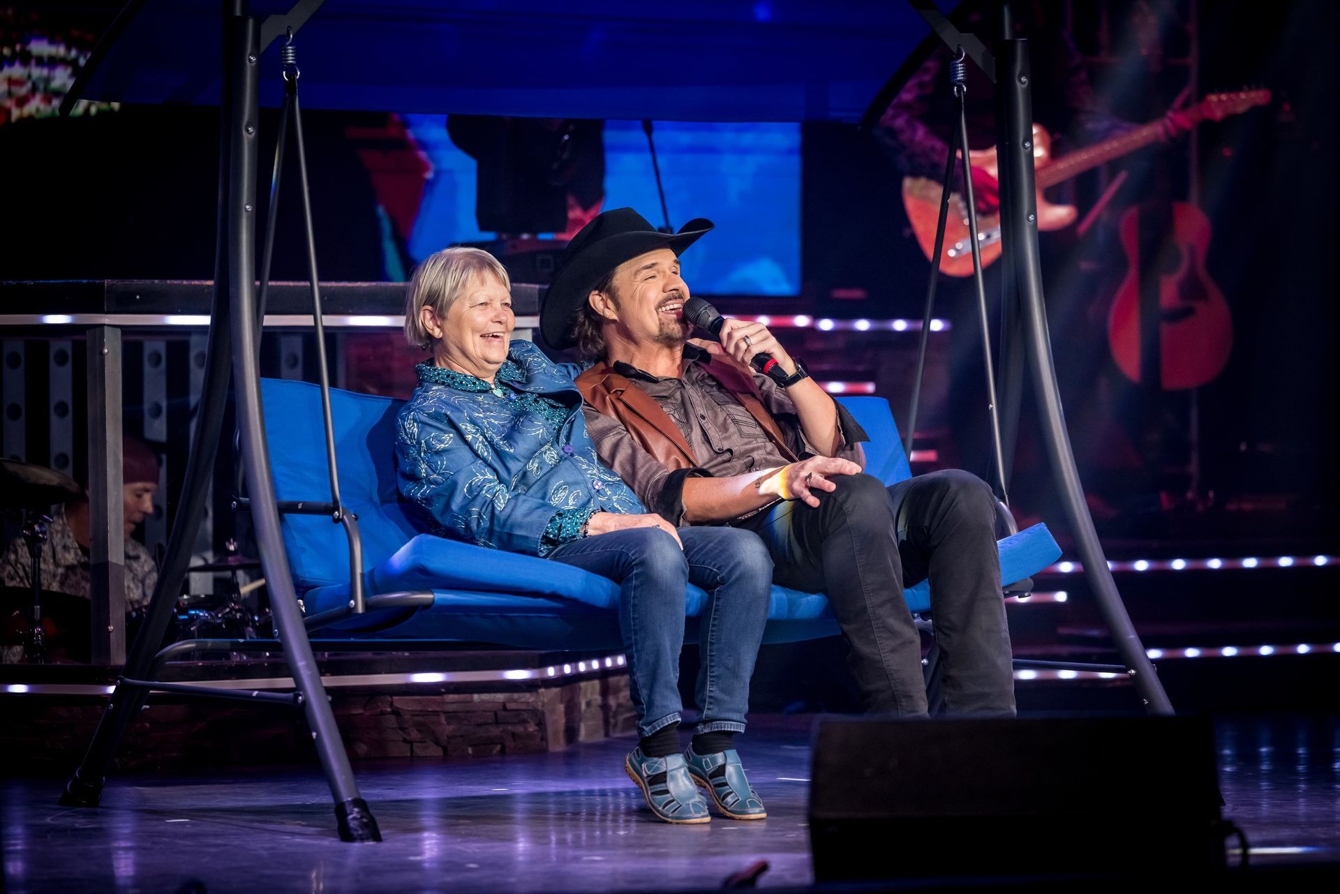 A person sits on a blue porch swing with a country singer holding a microphone on a stage with a guitar in the background.
