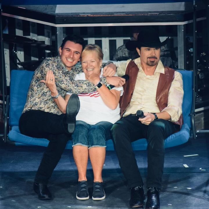 Two men and a woman sit together on a blue bench, smiling as the men embrace the woman.