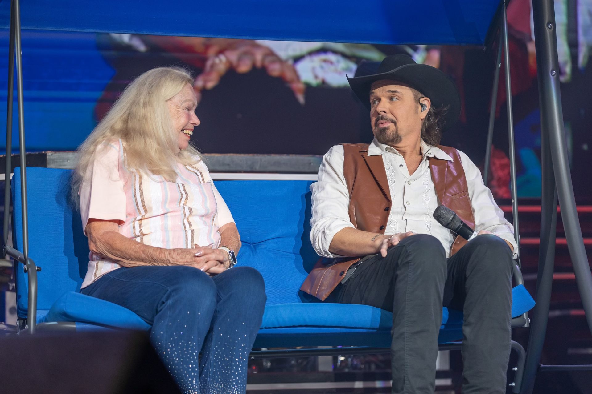 A blonde person and a person in a cowboy hat sit together on a blue swing bench, smiling and talking on stage.