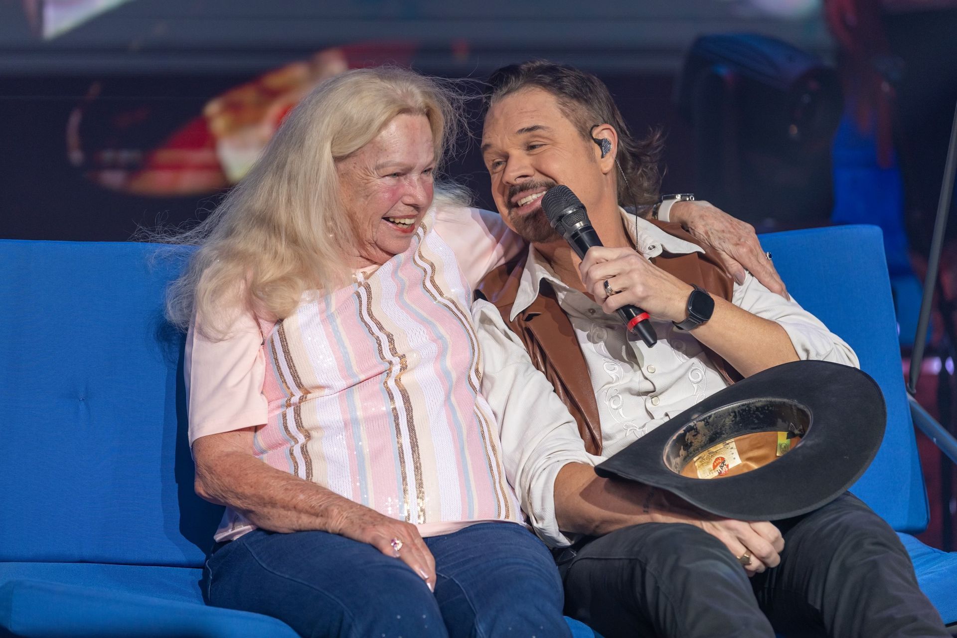 A smiling person in a western-style vest sits on a blue couch, embracing an older person while holding a microphone.