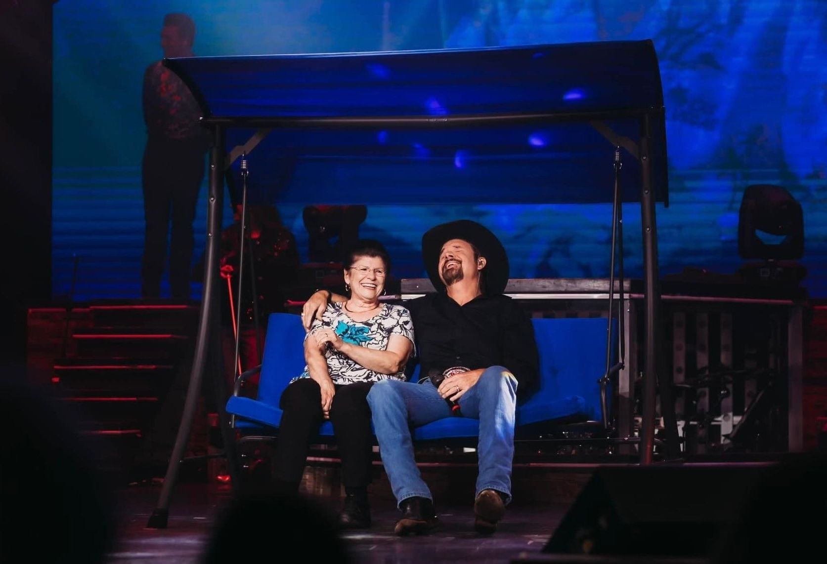 Two people sit smiling on a blue porch swing on a dark stage with a blue-lit backdrop.