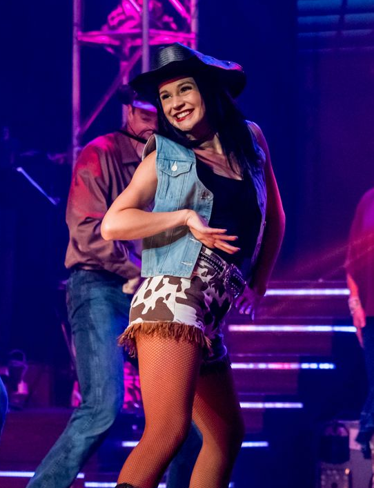 A young woman with dark hair wearing a black cowboy hat and cow printed shorts dancing with her hands on her hips 