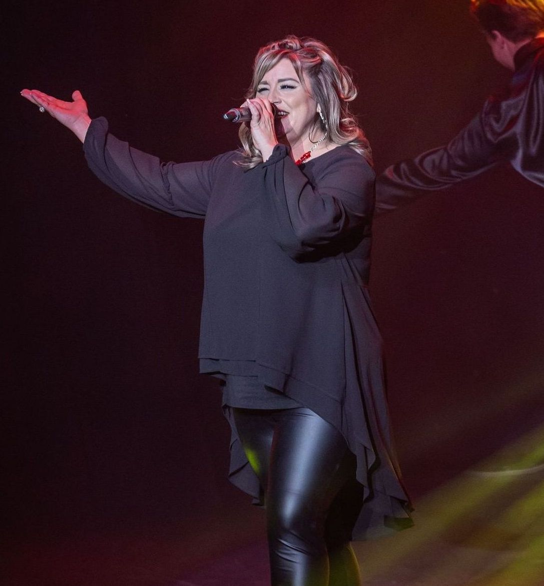 A person sings into a microphone on stage, wearing a black long-sleeved tunic and leather pants with one arm outstretched.