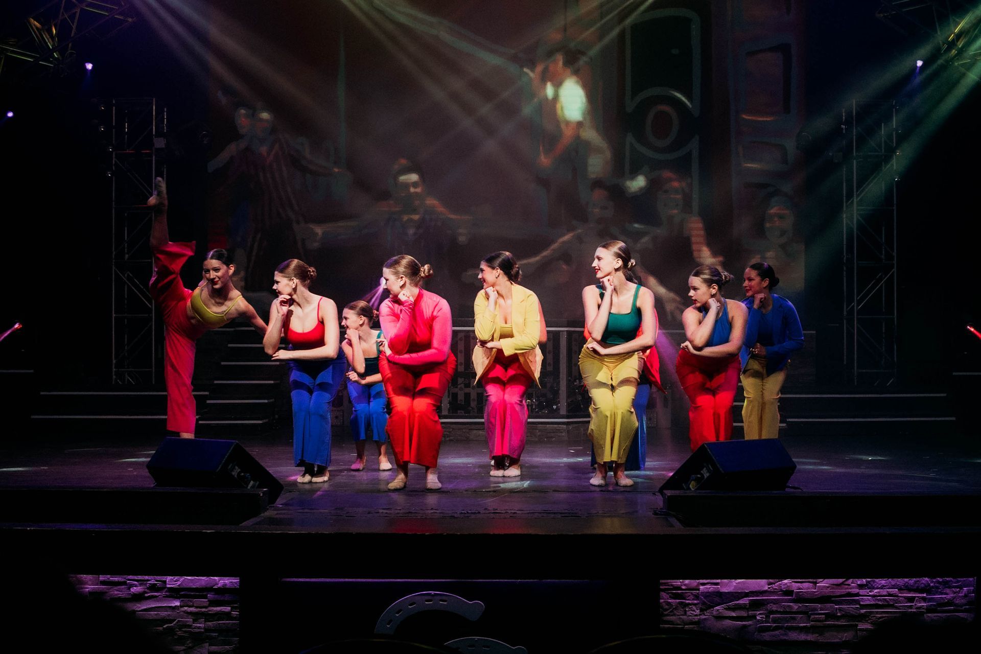 Dancers in colorful outfits perform a choreographed routine on a theater stage under spotlights.