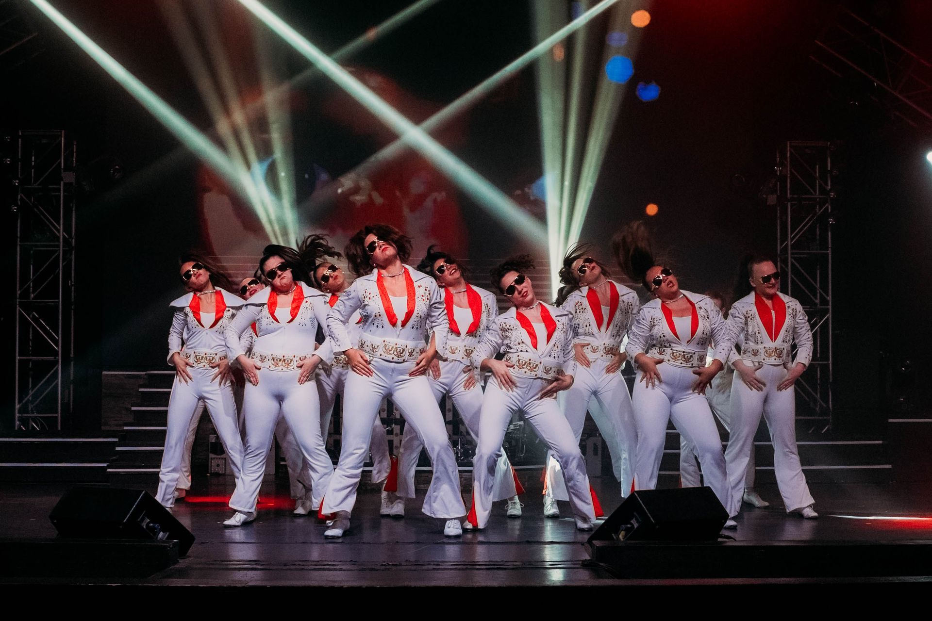 A group of dancers in white Elvis-style jumpsuits with red accents performing on a stage under bright spotlights.