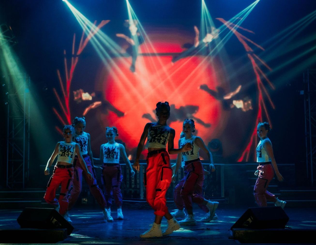 A group of performers dancing on a stage bathed in bright spotlights against a vibrant red backdrop.