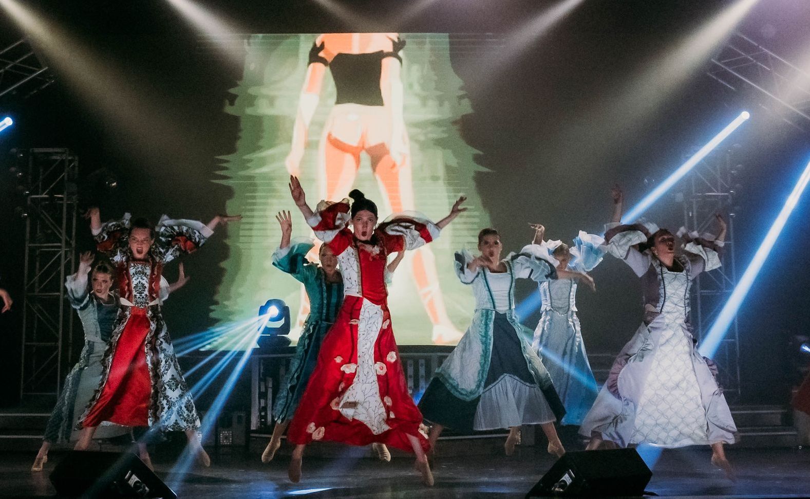 Performers in elaborate, flowing gowns dance on a stage with dramatic lighting and a large screen background.