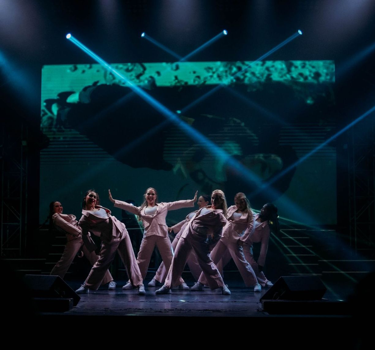 A group of dancers in light-colored suits perform in a dimly lit theater, illuminated by beams of blue stage lighting.
