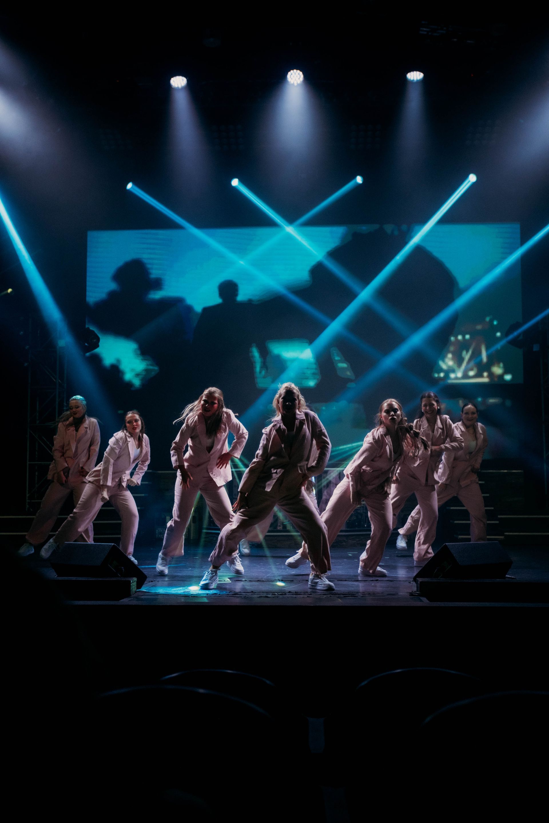 A group of dancers in matching light-colored outfits performs on a stage under bright, dramatic blue spotlights.