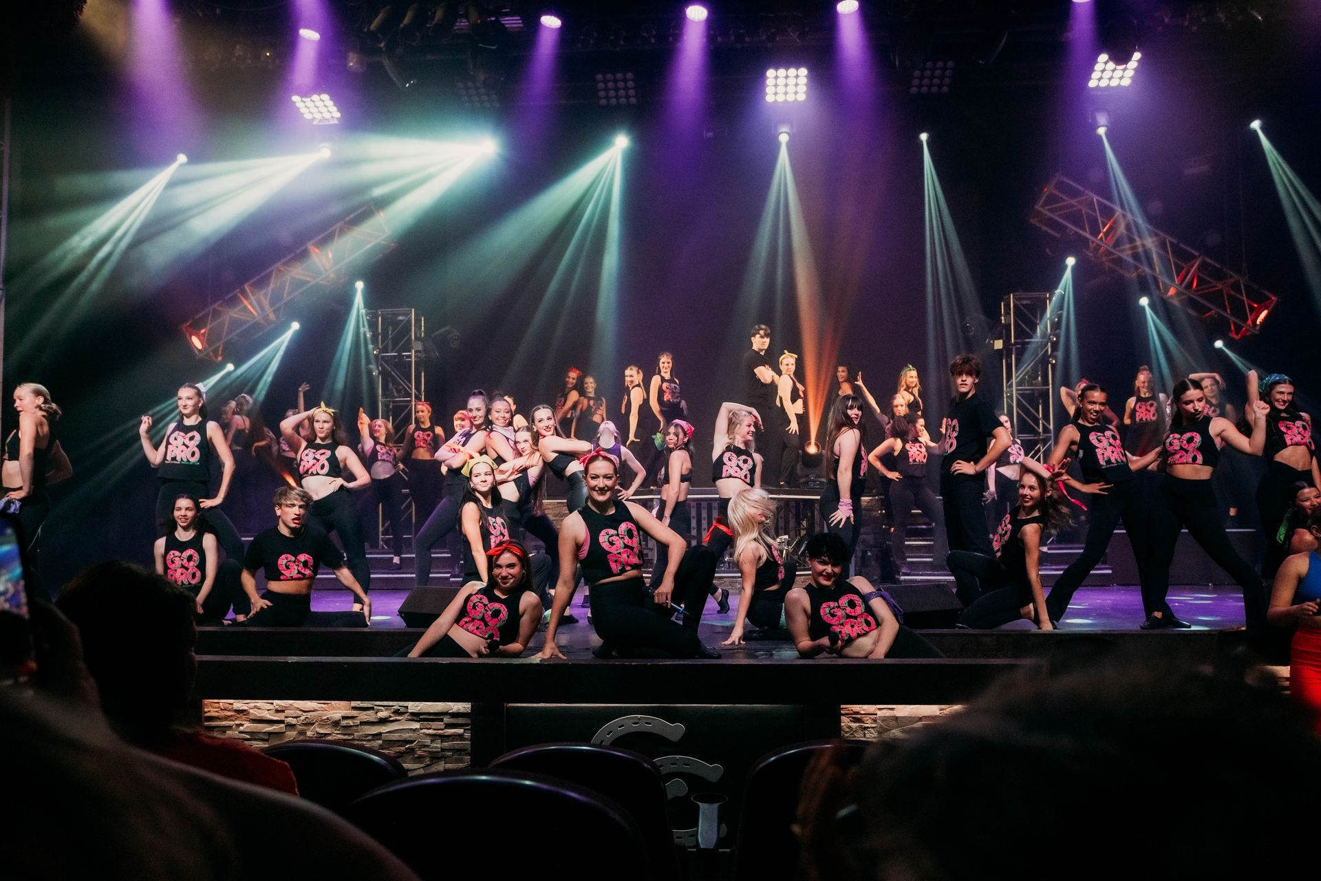 A large group of dancers in black stage outfits perform under bright, multi-colored stage lights.