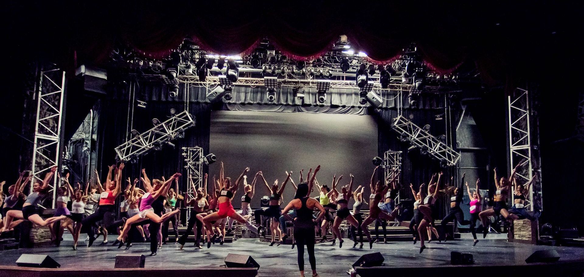 A group of dancers performs a synchronized routine on a brightly lit stage under metallic truss lighting rigs.