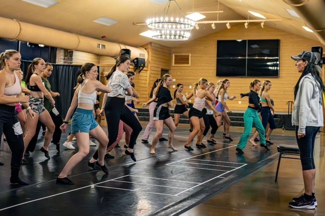 A group of dancers practices choreography in a wood-paneled studio while an instructor observes from the side.