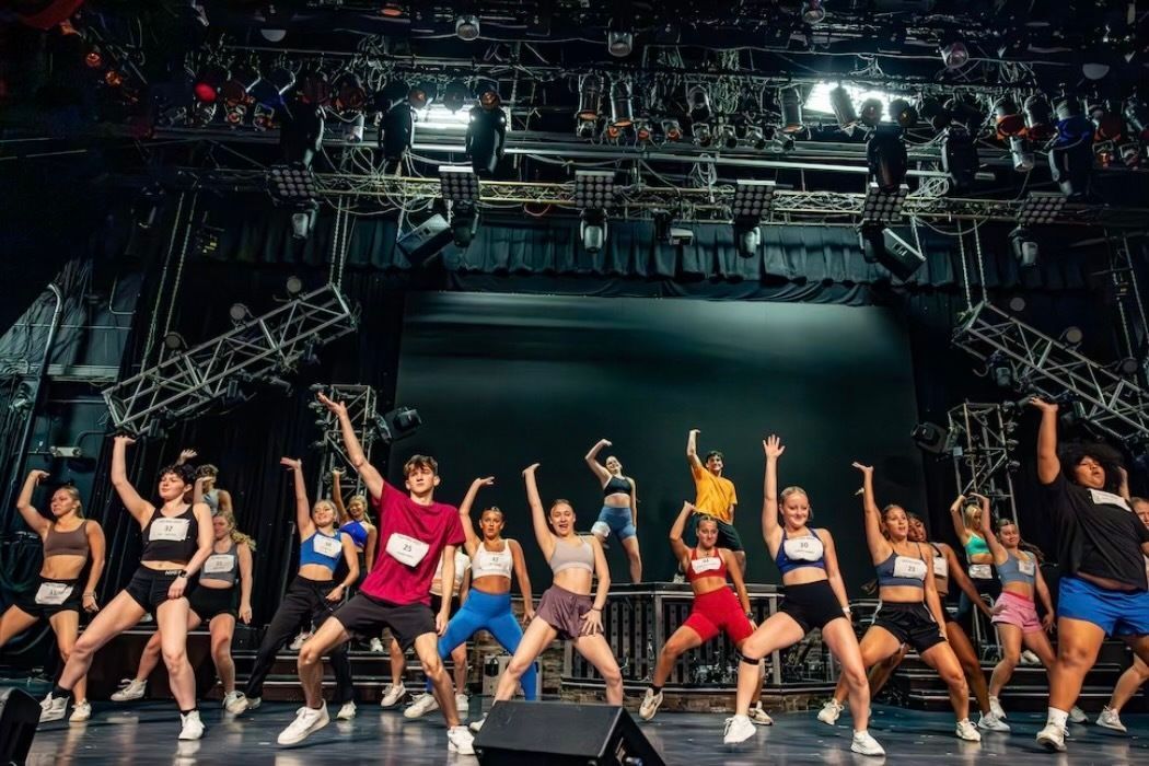 A group of people perform a coordinated dance routine on a stage with overhead lighting and stage rigging.