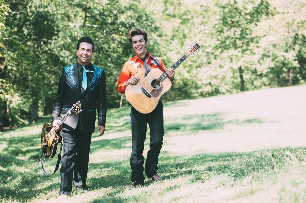 Two musicians walking on grass, carrying saxophone and guitar, with trees in the background.
