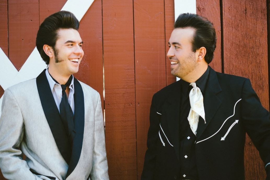 Two men in vintage-style suits smiling at each other against a red wooden wall.
