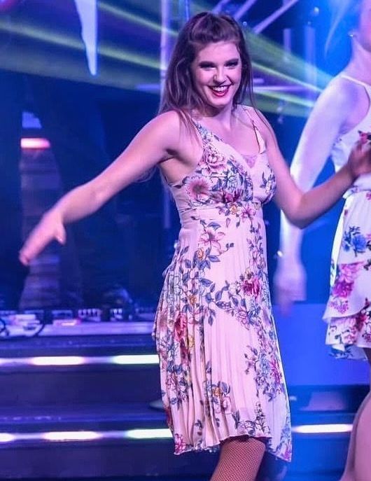 A person with a bright smile dances on a stage, wearing a light-colored dress with a colorful floral pattern.