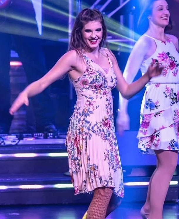 Two performers dance on stage wearing floral dresses, smiling under blue stage lighting.
