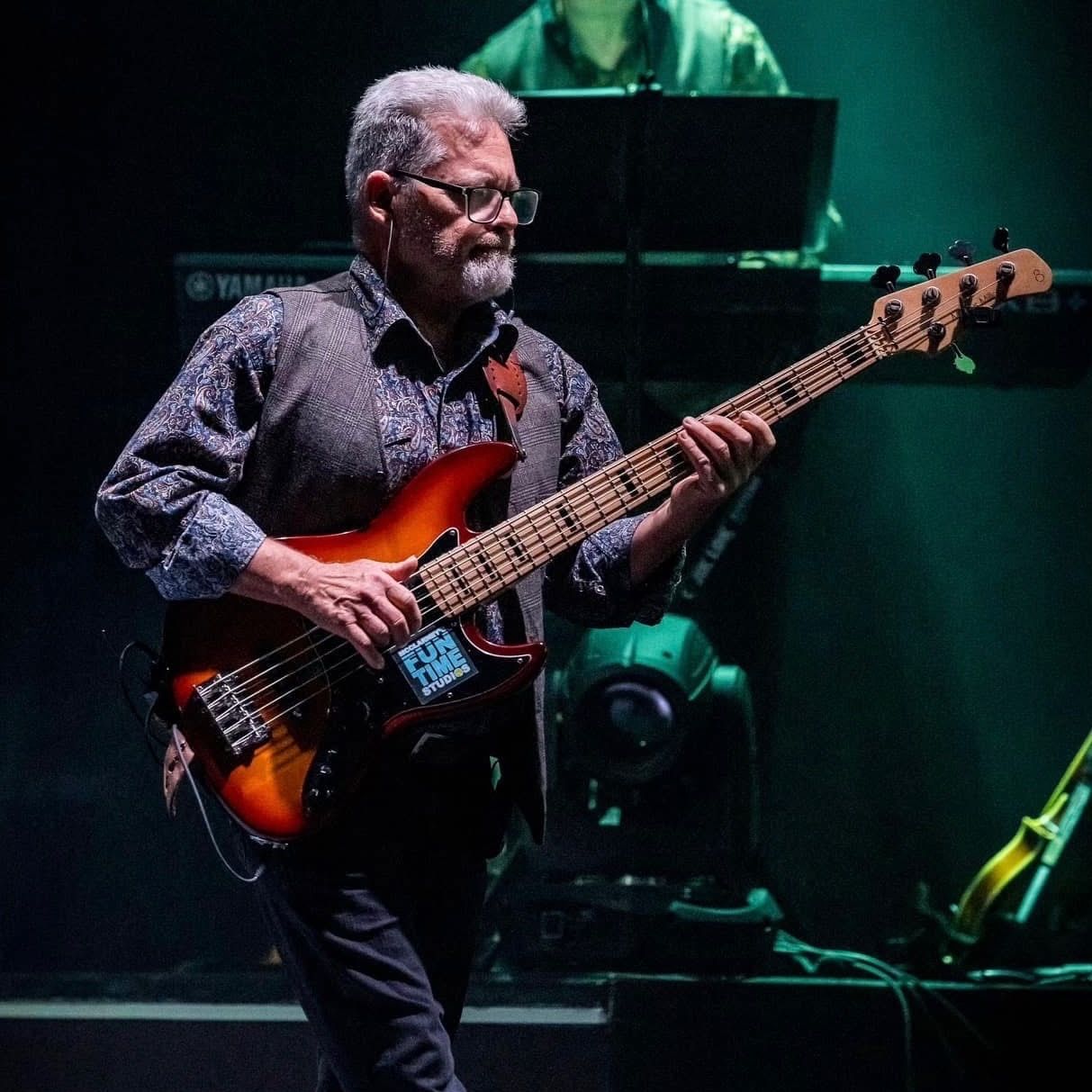 A bassist with glasses playing a sunburst five-string electric bass on a dimly lit stage.