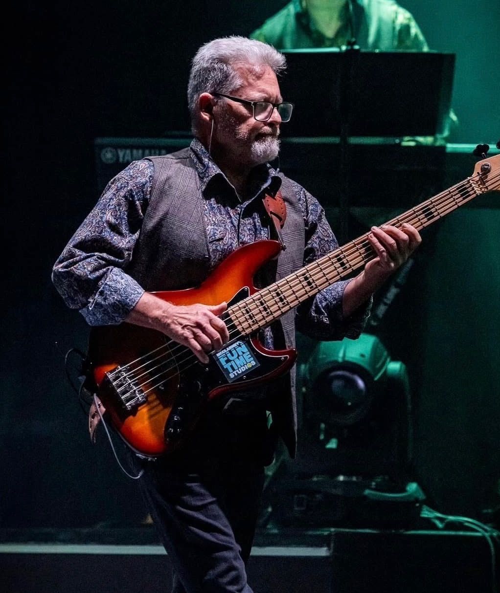 A musician with glasses and a patterned shirt plays a sunburst electric bass guitar on a dark stage under green light.