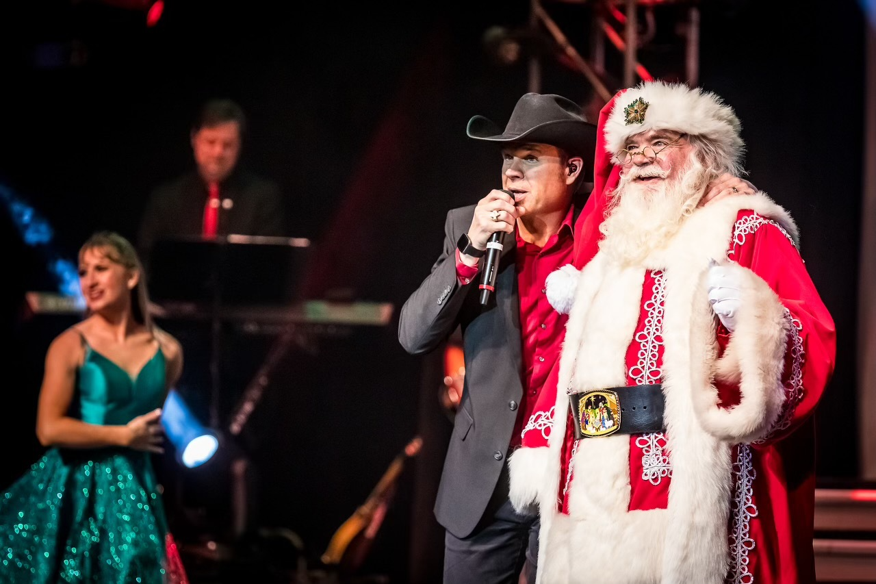 A person in a cowboy hat singing into a microphone next to Santa Claus, with a performer in a green dress nearby.