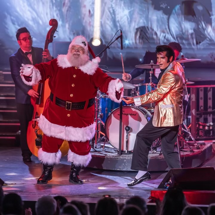 Santa Claus and an Elvis Presley impersonator in a gold jacket perform on a concert stage with a band.